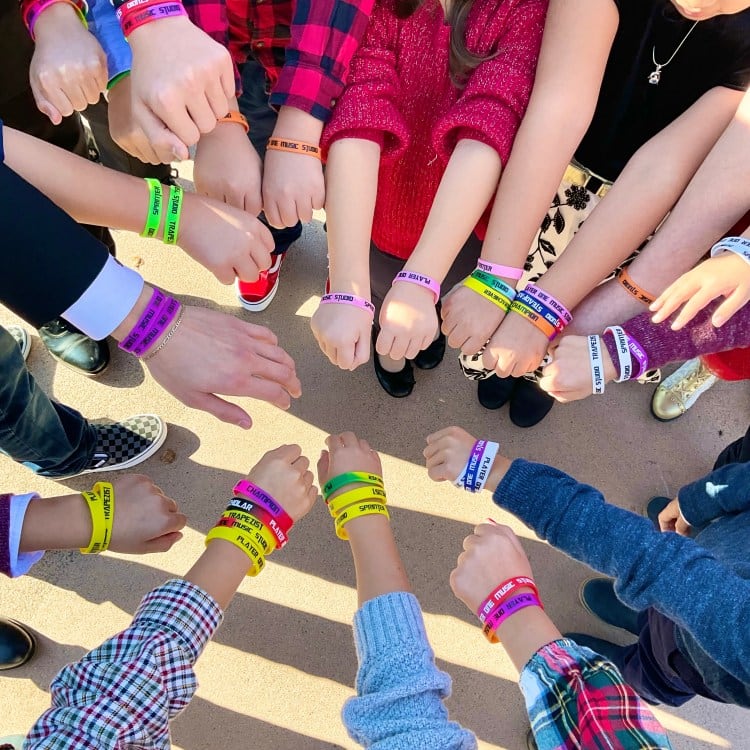 A ring of students' forearms, each wrist stacked with the printed Player One Music Studio bands they have earned.