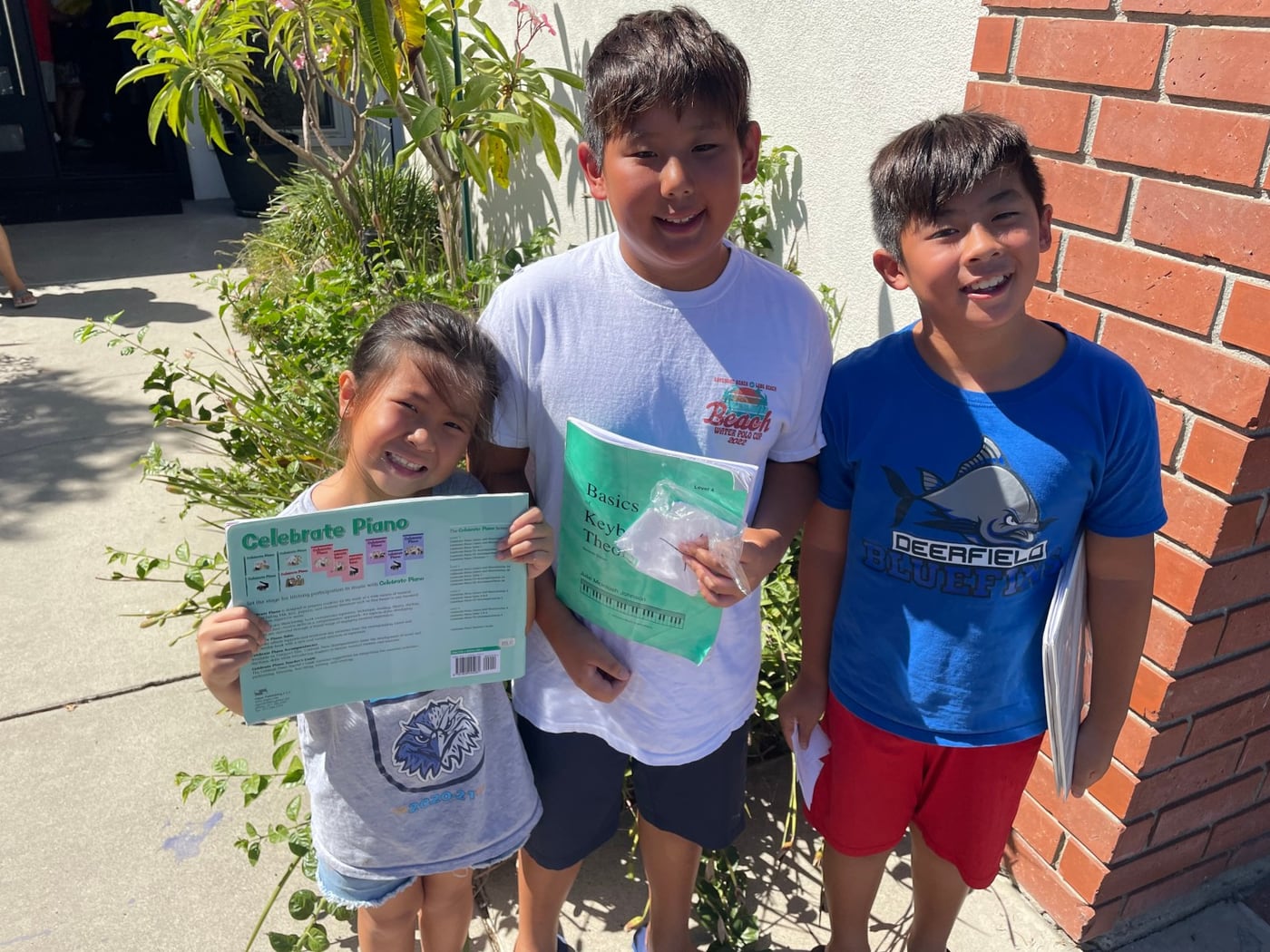 Three young students standing outside holding up the piano books they finished.