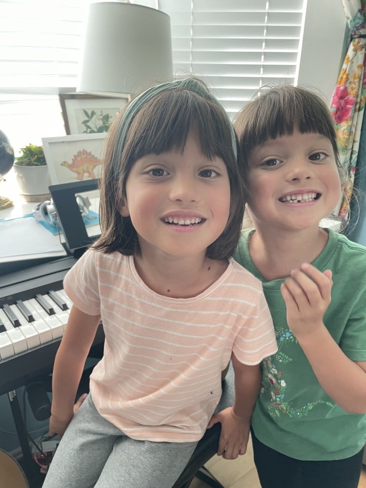 Two young students perched together on the bench at an upright piano, both smiling.